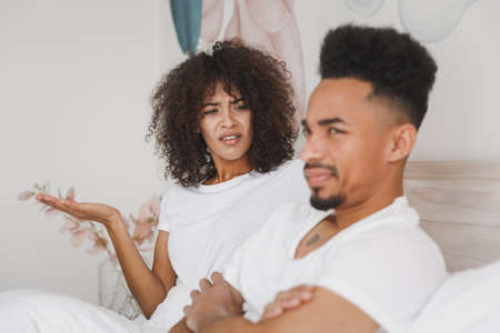 Young angry african swearing screaming couple girl guy in t-shirts lying on bed with white sheet pillow blanket indoors in bedroom at home spending time in room. Bad mood quarantine lifestyle concept.の写真素材