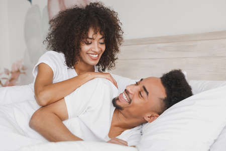 Young calm african loving couple girl guy in t-shirts lying on bed with white sheet pillow blanket indoors in bedroom at home, spending time in room. Rest relax good mood quarantine lifestyle concept.の写真素材