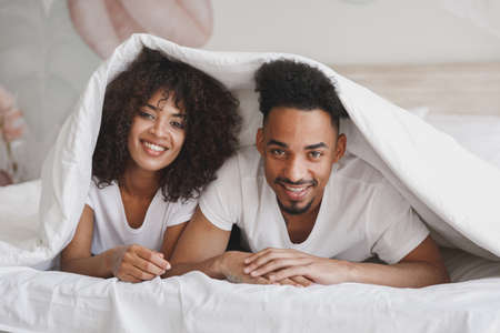 Young calm african loving couple girl guy in t-shirts lying on bed with white sheet pillow blanket indoors in bedroom at home, spending time in room. Rest relax good mood quarantine lifestyle concept.の写真素材