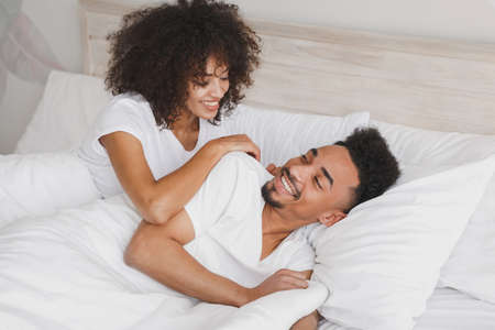 Young calm african loving couple girl guy in t-shirts lying on bed with white sheet pillow blanket indoors in bedroom at home, spending time in room. Rest relax good mood quarantine lifestyle concept.の写真素材