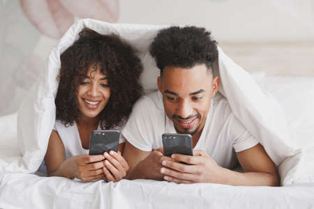 Young calm african loving couple girl guy in t-shirts using mobile cell phone lying on bed with...