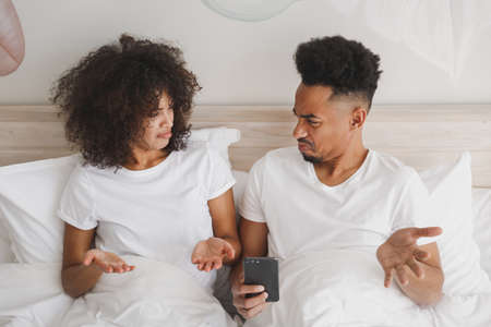 Young sad upset african couple girl guy in t-shirts using mobile cell phone lying on bed with white sheet pillow blanket indoors in bedroom at home, spend time in room. Quarantine lifestyle concept.の写真素材
