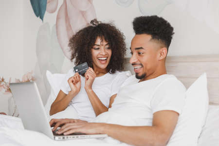 Young calm african loving couple girl guy in white t-shirts using laptop pc credit bank card lying on bed in bedroom at home, spending time in room. Rest relax good mood quarantine lifestyle concept.の写真素材