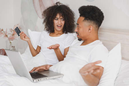 Young irritated frustrated shocked african couple girl guy in white t-shirts using laptop pc credit bank card lying on bed in bedroom at home spend time in room. Quarantine fake news lifestyle conceptの写真素材