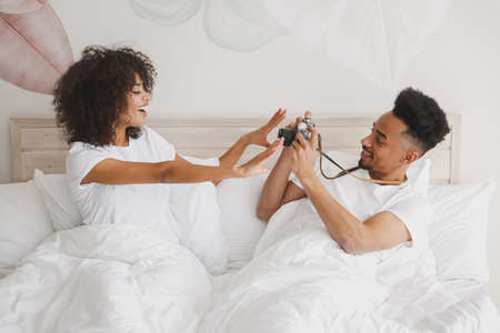Young african loving couple girl guy in white t-shirts lying on bed doing shot on retro vintage photo camera in bedroom at home, spend time in room. Rest relax good mood quarantine lifestyle concept.の写真素材