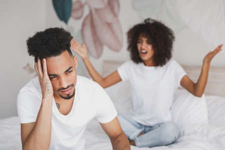 Young angry sad irritated frustrated african swearing screaming couple girl guy in white t-shirts sitting on bed indoors in bedroom at home spending time in room. Bad mood quarantine lifestyle conceptの写真素材