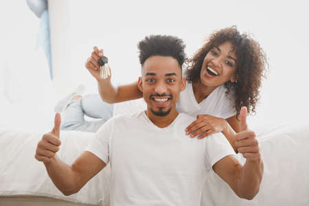 Young happy african loving couple girl guy in white t-shirts lying on bed hold in hand key from house indoors in bedroom at home, spend time in room. Rest relax good mood quarantine lifestyle concept.の写真素材