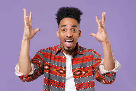 Shocked young african american guy in casual colorful shirt posing isolated on violet wall...
