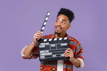 Smiling young african american guy in casual colorful shirt posing isolated on violet wall background studio. People lifestyle concept. Mock up copy space. Hold classic black film making clapperboard.の写真素材