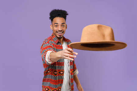Smiling young african american guy in casual colorful shirt posing isolated on violet wall background studio portrait. People sincere emotions lifestyle concept. Mock up copy space. Throwing out hat.の写真素材