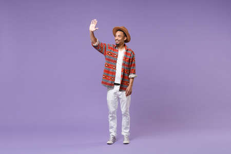 Smiling young african american guy in casual colorful shirt hat posing isolated on violet background. People lifestyle concept. Mock up copy space. Waving and greeting with hand as notices someone.の写真素材