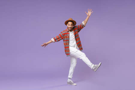 Excited young african american guy in casual colorful shirt hat posing isolated on violet background studio portrait. People lifestyle concept. Mock up copy space. Dancing rising leg, spreading hands.の写真素材