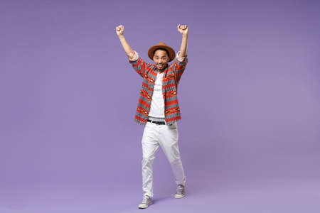 Joyful young african american guy in casual colorful shirt hat posing isolated on violet wall...