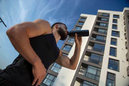 Exhausted athlete after exercise drinks water from a black bottle. White high-rise on the...