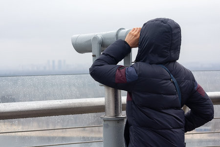 A tourist looks through a telescope at a panorama of the city in rainy weather
