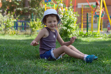 A child in a panama hat sits on the lawn near the playground. Peopleの写真素材