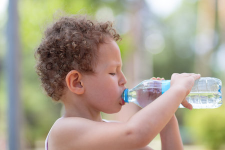 A curly child drinks water from a plastic bottle on a hot day. Peopleの写真素材