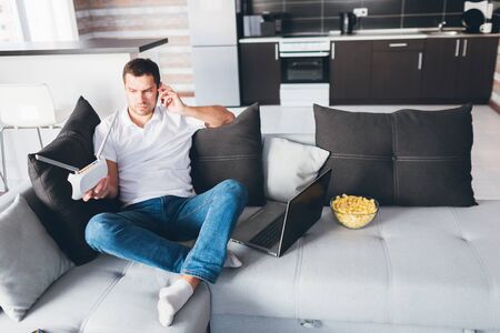 Young man watch tv in his own apartment. Guy sit on couch and talking on phone. Hold wi-fi router in hand and look at it. Customising internet device. Opened laptop on couch besides.の写真素材