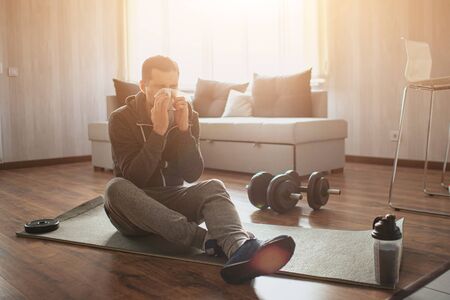 Young ordinary man go in for sport at home. Picture of spot beginner sitting on mat and sneezing. Sick and ill. Caught flu. Suffer from pain. Freshman in sport after or before workout got sick.の写真素材