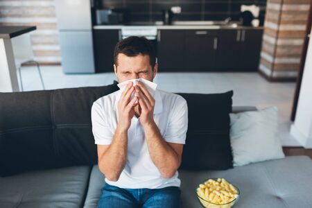 Young man watch tv in his own apartment. Guy sneezing into white napkin. Sick and ill at home. Suffer from headache or nose pain. Serious disease and sickness. Sit alone at home.の写真素材