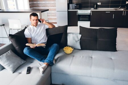 Young man watch tv in his own apartment. Serious concentrated guy reading book and talking on phone at same time. Hold opened laptop on knees. Studying or working. Remote out of office.の写真素材