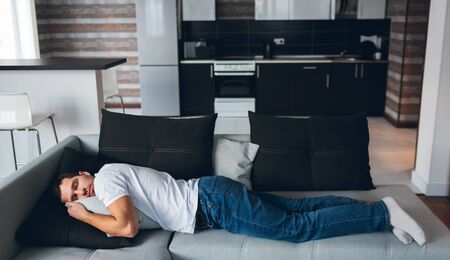 Young man watch tv in his own apartment. Guy lying in horizontal position on couch. Daydreaming with...