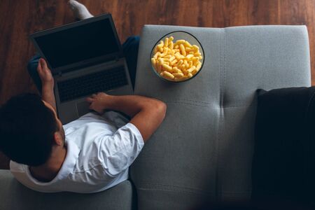 Young man watch tv in his own apartment. Sit alone on floor with legs crossed and hold laptop on it. Watching movie, sports program. Entertain himself. Up view.の写真素材