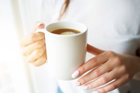 Young woman on kitchen during quarantine. Stand at window with curtains and hold white cup of tea or coffee. Close up.の写真素材