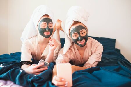 Two female models make face mask and using smartphone or phone for selfie and communication in social networks. Two young women in towels and pajamas have a fun spa party together in home.の写真素材