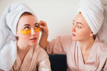 Two young women having fun with patches under their eyes. Two friends in towels and pajamas have a fun spa party together in home.の写真素材