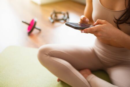 Young woman doing yoga workout in room during quarantine. Cut view of girl using smartphone. Woman sit on mat during rest.の写真素材