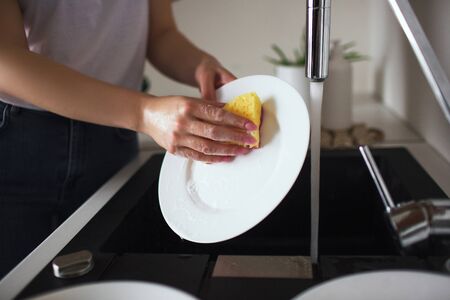 Young woman in kitchen during quarantine. Whshing white plates with sponge and dishwasher. Cleaning dishes alone in kitchen. Cut view.の写真素材