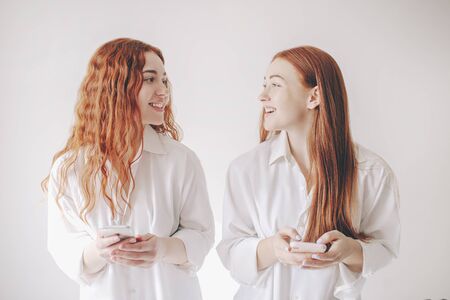Two red-haired sisters stand isolated on a white background in spacious oversized shirts. Two young women are addicted to social networks spend all their time in a smartphoneの写真素材