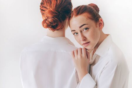 Two red-haired sisters stand isolated on a white background in shirts. One woman show their backs....