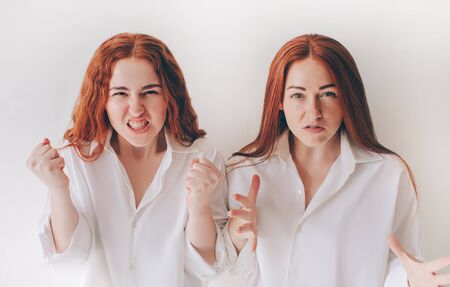 Two young women got angry and shout at the camera They feel rage, aggression, anger. Two red-haired sisters stand isolated on a white background in spacious oversized shirtsの写真素材