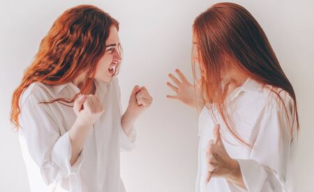 Two red-haired sisters stand isolated on a white background in spacious oversized shirts. Two young women got angry and shout at each other They feel rage, aggression, angerの写真素材