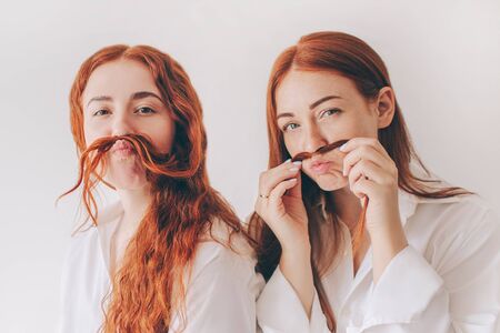 Two red-haired sisters stand isolated on a white background in spacious oversized shirts. Two young...