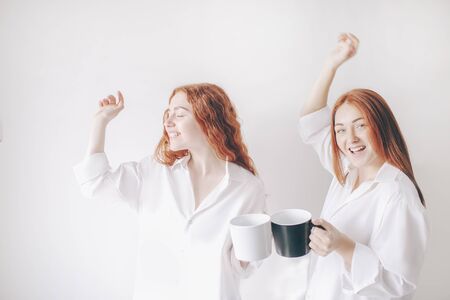 Two red-haired sisters stand isolated on a white background in spacious oversized shirts. Two young girls drink coffee or tea and fool around and have funの写真素材