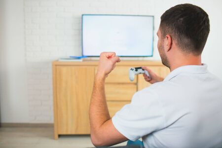 Young man sit in room during quarantine. Guy playing tv computer game using joystick. Cheering alone...