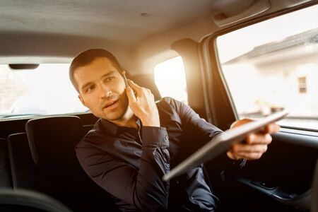 Busy businessman in a taxi. Multitasking concept. Passenger rides in the back seat and works simultaneously. Speaks on a smartphone and uses a tablet pc and communicates with the driverの写真素材