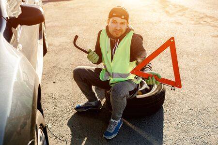 Worker or driver repairing a car. He throws up his hands and does not know what to do. Concerned and worried. Bad worker. He is dressed in a signal vest. He has a road triangle in his hands. Sits on a bus from a carの写真素材
