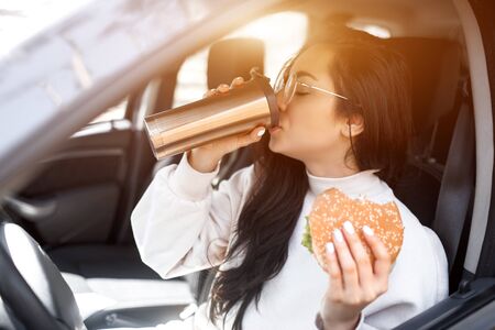 Beautiful brunette model has lunch in a car. Woman eats a hamburger and drinks coffee or tea . Food...