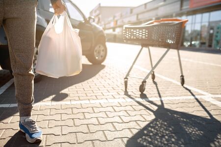 Close-up of a stroller with food near a large supermarket in a suburban shopping center. A man stands near a car in a parking lot after a successful shoppingの写真素材