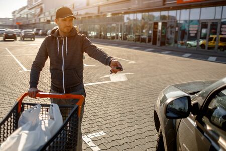 Close-up of a stroller with food near a large supermarket in a suburban shopping center. A man stands near a car in a parking lot after a successful shoppingの写真素材