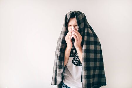 Young sick man isolated over white background. Guy covered with blanket. Sneezing into white tissue...