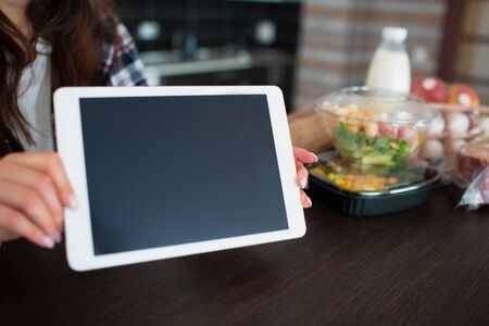 Food delivery concept. A young woman orders food using a laptop at home. On the table are milk,...