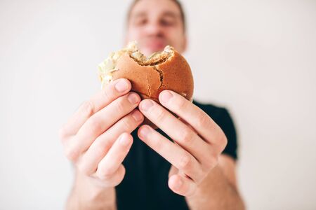 Young man isolated over white background. Guy hold bited cheeseburger in hands and show it on camera. Defocused or blurred background. Satisfied happy guy on picture.の写真素材