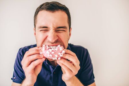 Young man isolated over background. Guy biting pink donut piece with pleasure. Hungry person divour...