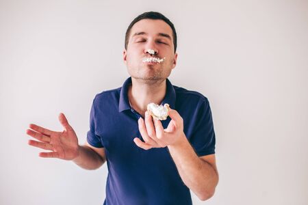Young man isolated over white background. Eating tasty delicious fat small cake. Part of cream is on his nose and face. Yummy pleasure.の写真素材