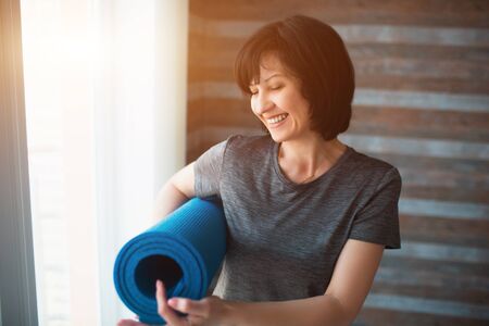 Adult fit slim woman has workout at home. Cheerful positive senior female person hold yoga mat in hand and point on it. Smiling during standing in room before workout.の写真素材